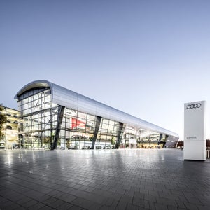 A modern glass building with a courtyard, trees, and solar panels on the roof, with a blue sky in the background.