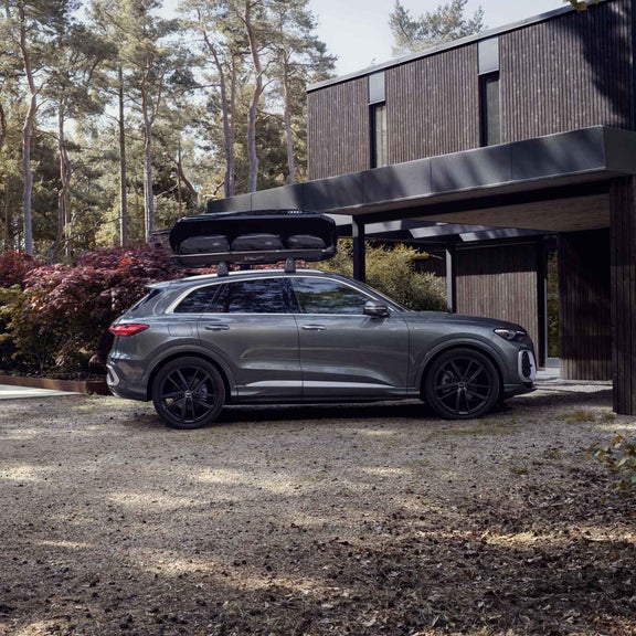 Der Q5 SUV ist mit einer Dachbox ausgestattet und steht seitlich vor einem Haus im Wald. 	