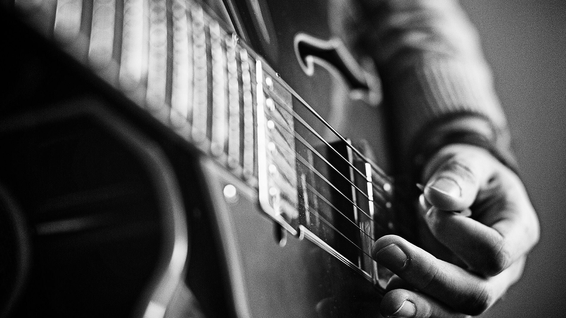 Nahaufnahme einer Hand, die Saiten einer E-Gitarre anschlägt; Bild in Schwarz-Weiß gehalten.