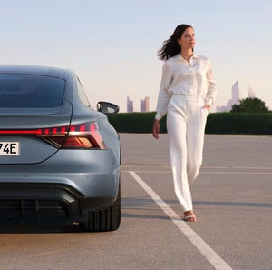 A woman in elegant white attire walks confidently beside a silver Audi car in an open parking lot, with a city skyline in the hazy background.