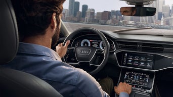 Mann fährt ein Audi mit digitalem Cockpit und Touchscreen, Skyline einer Stadt im Hintergrund durch die Frontscheibe sichtbar.
