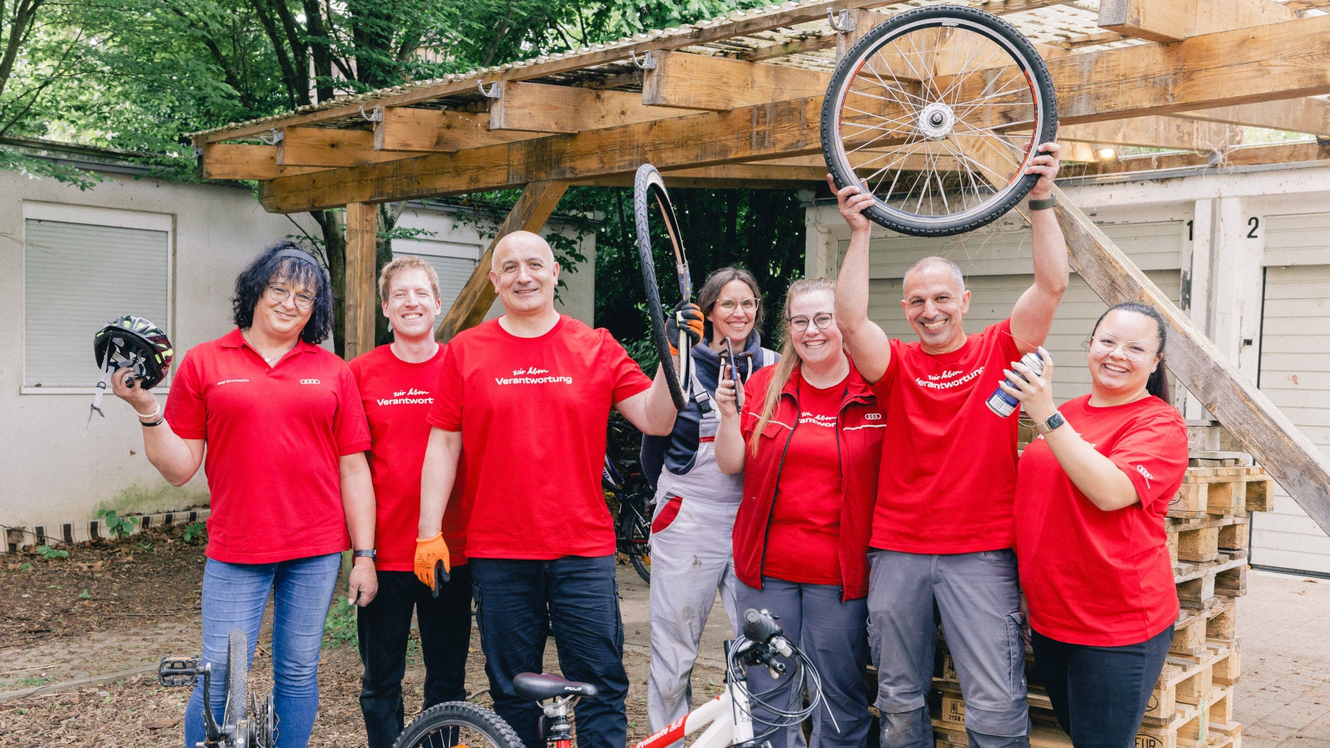 Mitarbeitende in roten T-Shirts stehen vor einer Garage und halten Fahrradteile in die Luft