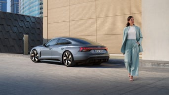 A woman in a stylish blue suit walks confidently beside a sleek grey Audi parked next to a modern building, conveying elegance and sophistication.