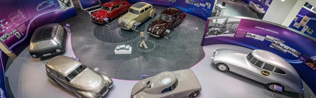 Vogelperspektive einer Oldtimer-Ausstellung mit sechs historischen Fahrzeugen in Rot, Beige, Dunkelrot und Silber; eine Person steht in der Mitte.
