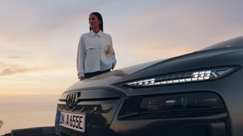 Szenische Außenaufnahme im Sonnenuntergang vor Küstenkulisse: im Vordergrund die Front eines Audi mit eingeschalteten Scheinwerfern. Dahinter steht eine dunkelhaarige Frau mit weißer Jacke.