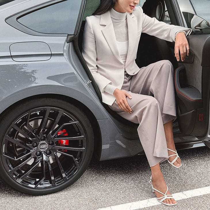Woman in a suit sitting in an open car door, sleek modern car wheel visible.