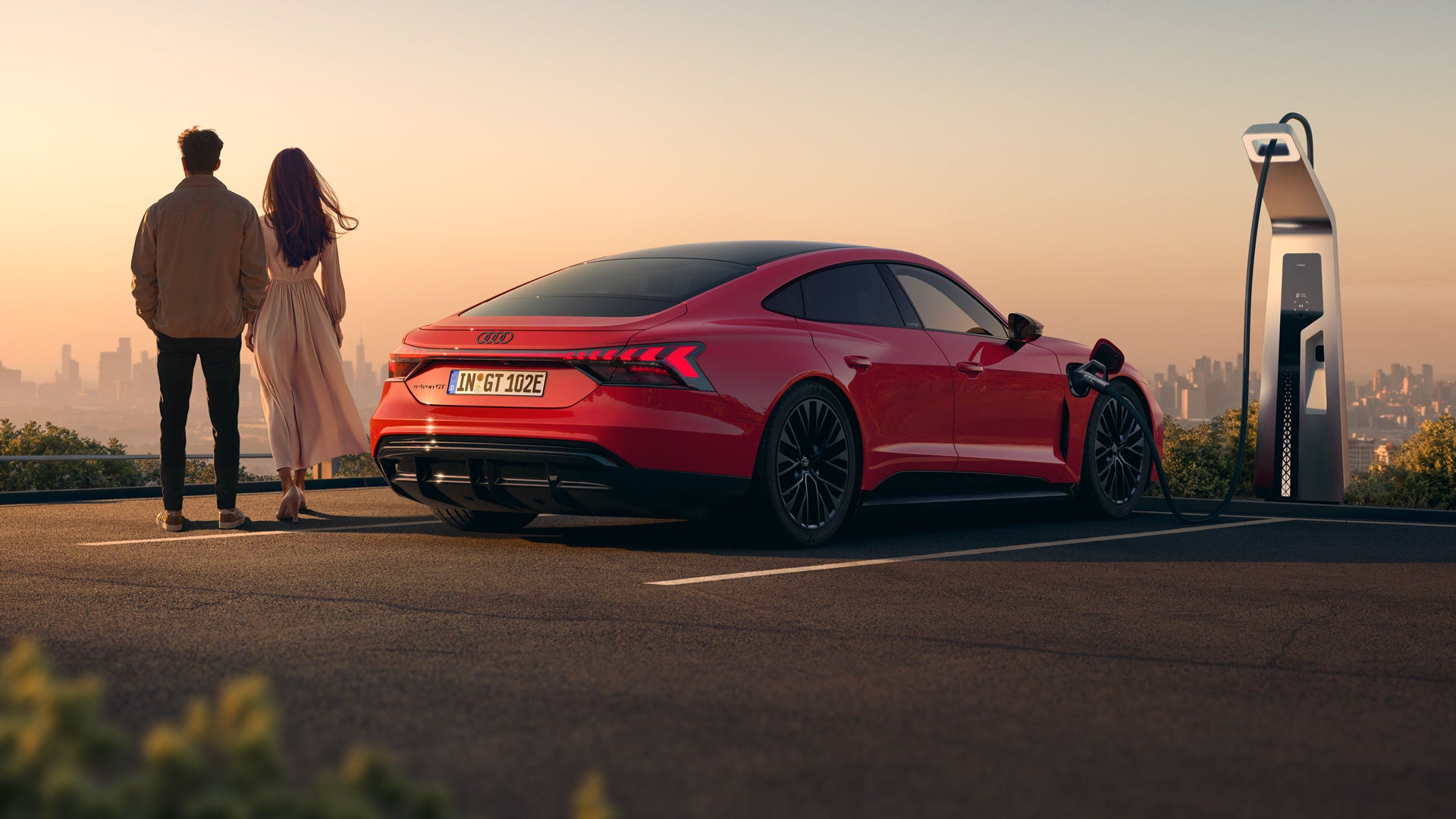 Ein roter Audi e-tron GT quattro steht auf einer Anhöhe über einer Stadt, die ins Licht eines Sonnenuntergangs gehüllt ist. Das Fahrzeug wird an einer öffentlicher Ladesäule geladen. Neben dem Fahrzeug stehen eine Frau und ein Mann.