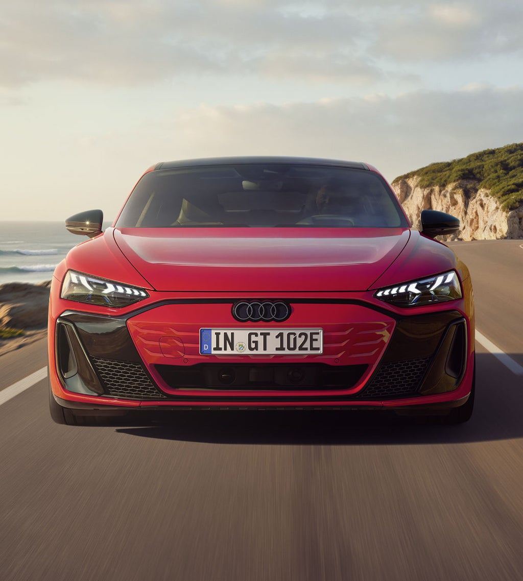 Frontansicht eines roten Audi e-tron GT quattro, der auf einer Küstenstraße fährt. Im Hintergrund sind Klippen, das Meer und der Himmel zu erkennen. Die gesamte Szenerie ist in das Licht eines Sonnenuntergangs gehüllt.