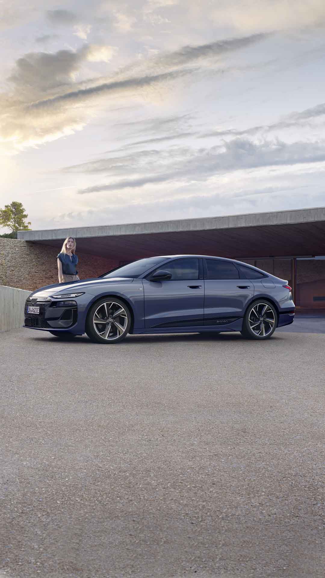 Der A6 Sportback e-tron steht im Seitenprofil vor dem Carport eines Hauses. Daneben steht eine junge Frau.