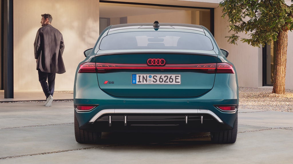 Heckansicht eines grünen Audi S6 Sportback e-tron mit leuchtendem Rücklicht, daneben ein gehender Mann im Mantel.