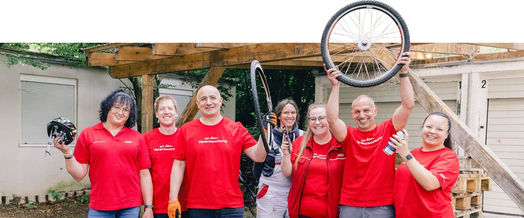 Mitarbeitende in roten T-Shirts stehen vor einer Garage und halten Fahrradteile in die Luft