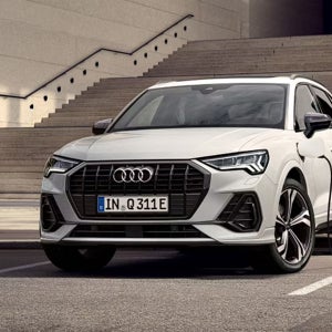 A grey Audi SUV is parked at a charging station beside a modern building. The scene suggests innovation and sustainability against a scenic backdrop.