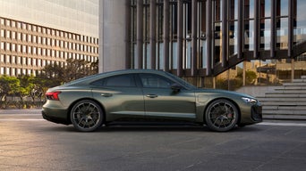 A green Audi RS e-tron GT Performance car parked on a city street, with modern buildings and greenery in the background under soft sunlight.