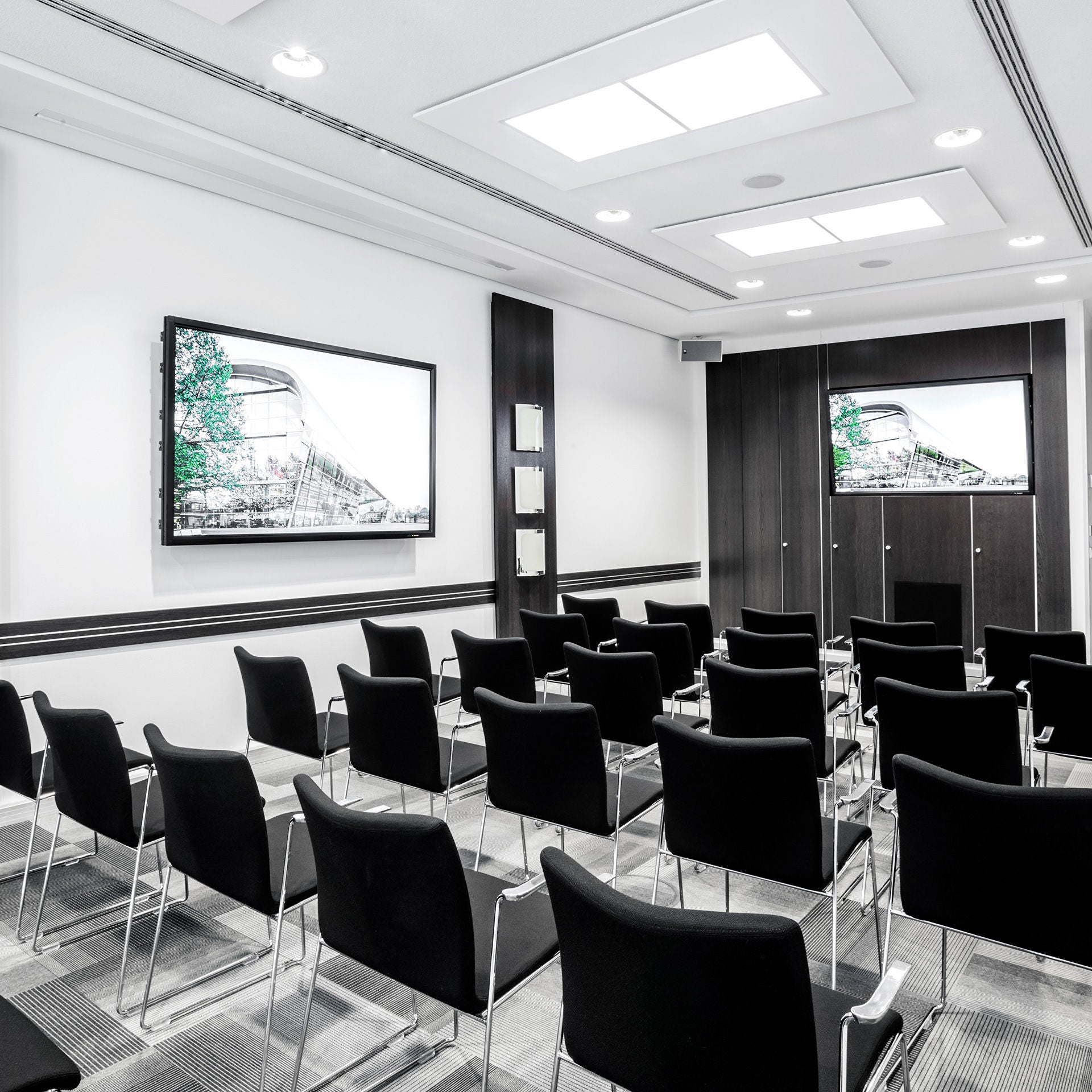 Bestuhlter Konferenzraum mit zwei Bildschirmen, Gebäudefotos und weißen Tischen.