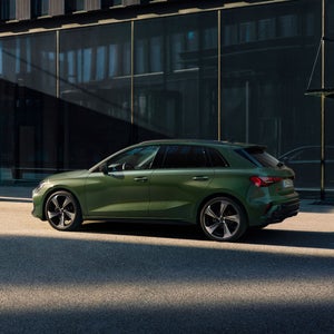 A green Audi A3 is parked on a sunlit city street, casting a shadow on the pavement. The modern building in the background adds a stylish, urban feel.