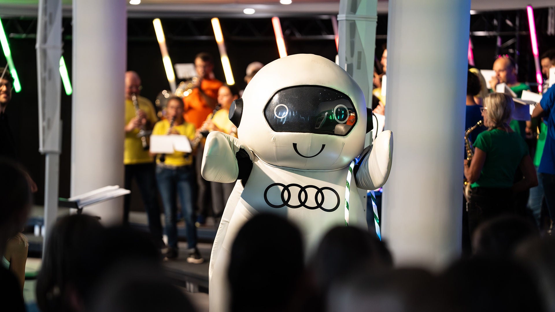 Weißes Audi Robotermaskottchen ADUI vor einem Orchester mit bunten Leuchtstäben bei einem Kinderkonzert.