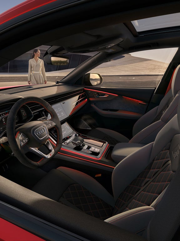Innenraum eines Audi SQ8 SUV mit Sportsitzen, roten Kontrastnähten und leuchtender Ambientebeleuchtung, Frau außen sichtbar.