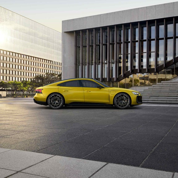 Gelber Audi RS e-tron GT performance geparkt vor einem modernen Gebäude mit Glasfassade und Treppe bei Sonnenuntergang. 