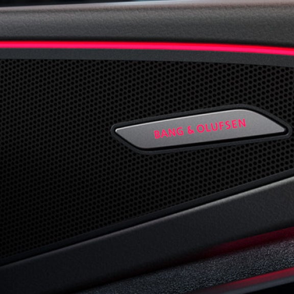 Close-up of a Bang & Olufsen car speaker, set in a black grid with sleek red ambient lighting.