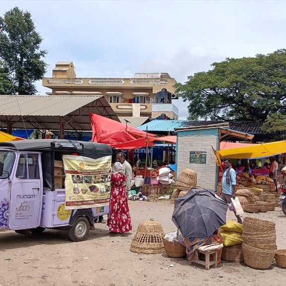Ein lilafarbenes E-Rikscha-Fahrzeug steht auf einem belebten Marktplatz mit Verkaufsständen und Passanten.