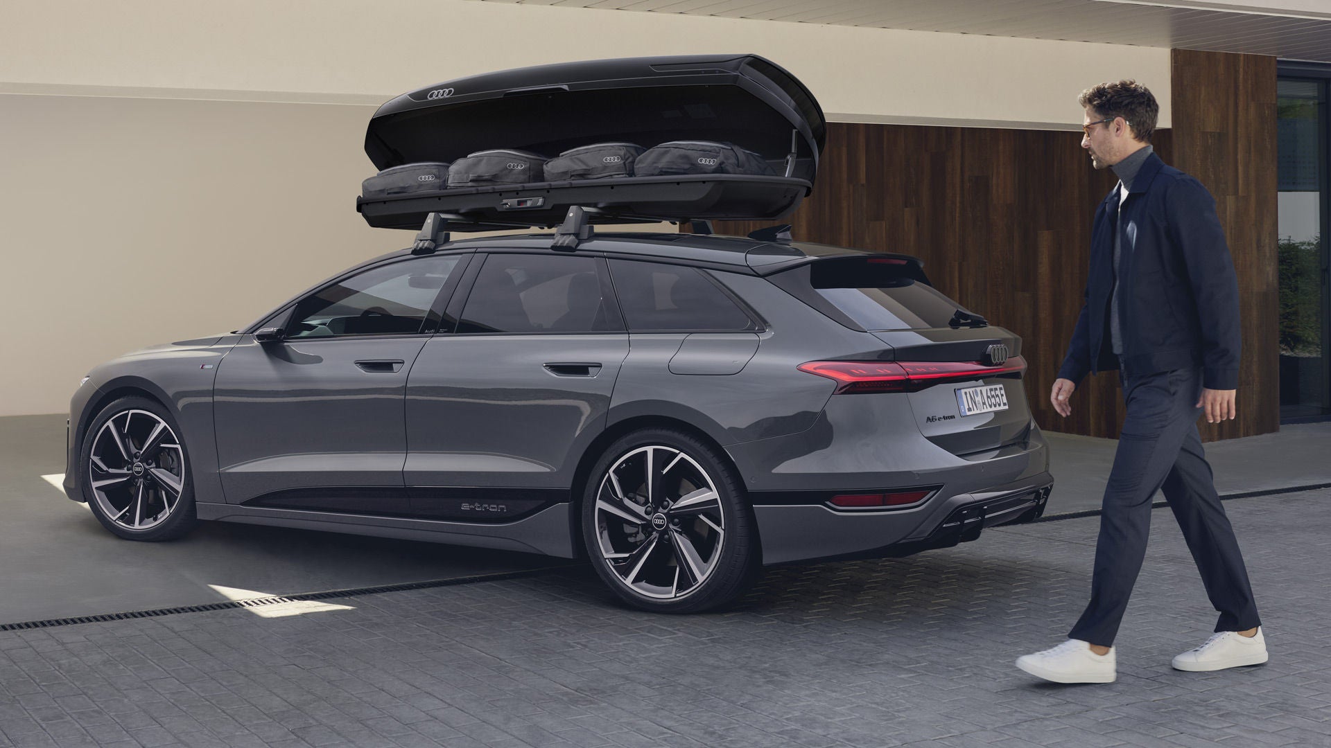 Der A6 Avant e-tron steht schräg vor einem modernen Haus, ein Mann bewegt sich auf das Fahrzeug zu. Auf dem Dach ist eine Dachbox montiert, deren Klappe geöffnet ist. Taschen sind in der Dachbox zu sehen.&nbsp;
