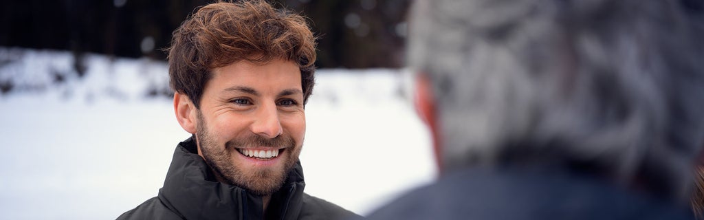 Lächelnder Mann mit braunen Locken im Winteroutfit im Gespräch mit einem grauhaarigen Mann im Vordergrund, verschneite Landschaft im Hintergrund.