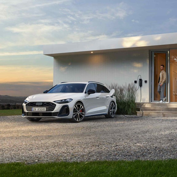 Ein weißer Audi A5 Avant e-hybrid beim Laden an einer Wallbox mit einen Mann der in der Tür steht