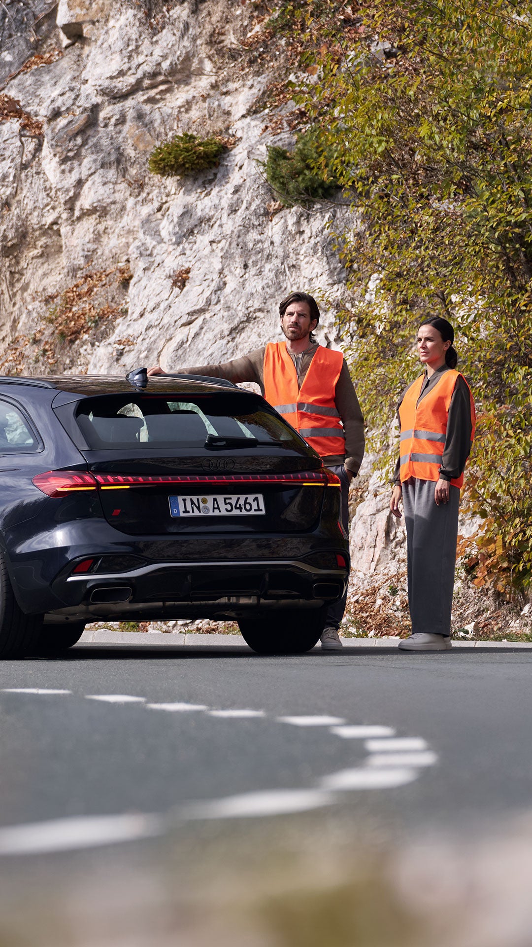 Ein Paar in orangen Warnwesten steht neben ihrem schwarzen Audi am Straßenrand einer Landstraße. Sie haben eine Panne und im Vordergrund sieht man ein rotes Warndreieck auf der Straße stehen.