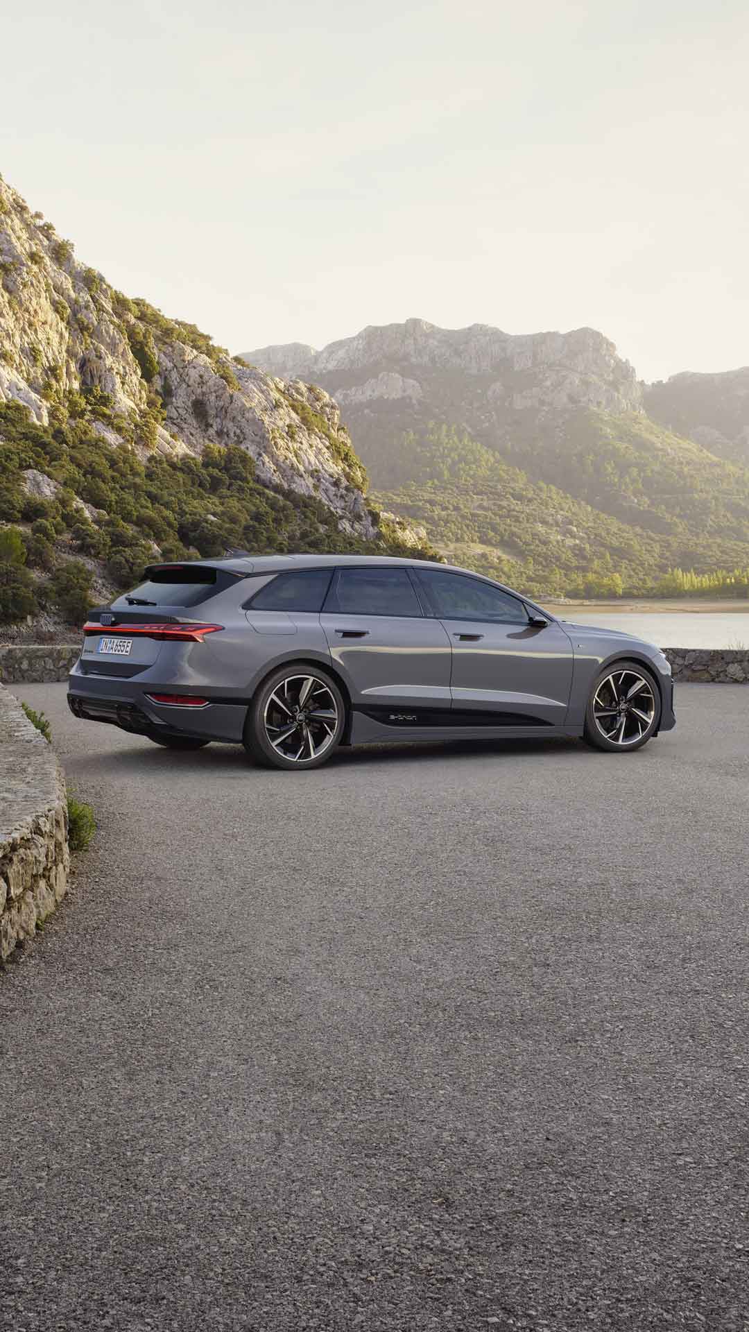 Der A6 Avant e-tron steht seitlich auf einem Kiesparkplatz vor einem See. Im Hintergrund sind von Bäumen bewachsene Berge zu sehen.&nbsp;