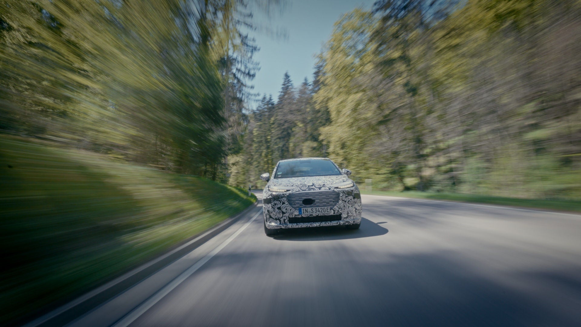 A camouflaged A2 e-tron drives down a country road through a green forest.