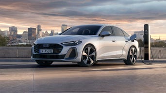 Eine weiße Audi A5 Limousine e-hybrid steht auf einem Parkdeck an einer Ladesäule. Im Hintergrund ist eine Großstadt zu sehen, die von der untergehenden Sonne angestrahlt wird.