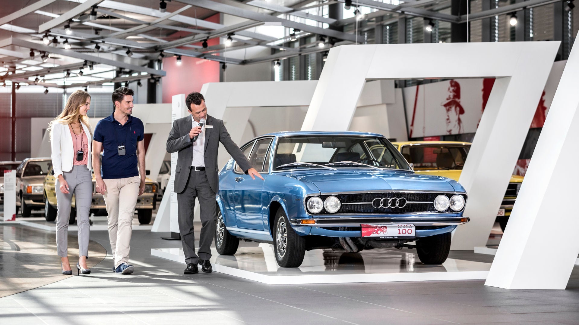 An employee shows two visitors a blue vintage Audi in an exhibition.