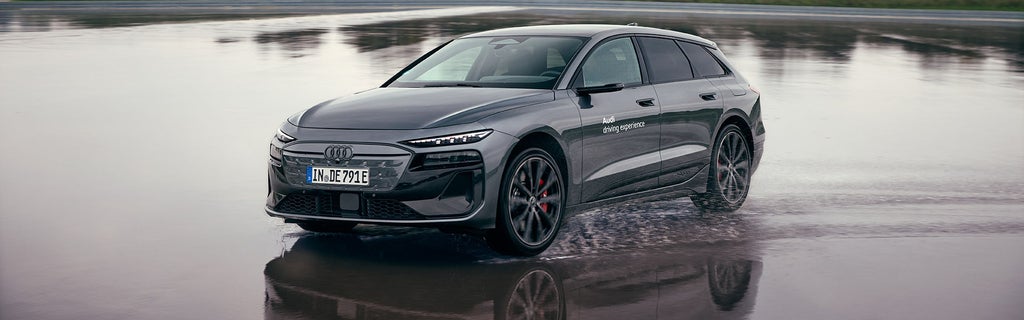 Ein grauer Audi S6 Avant e-tron mit der Aufschrift „Audi driving experience“ fährt über nasse Fahrbahn, Wasser spritzt auf.