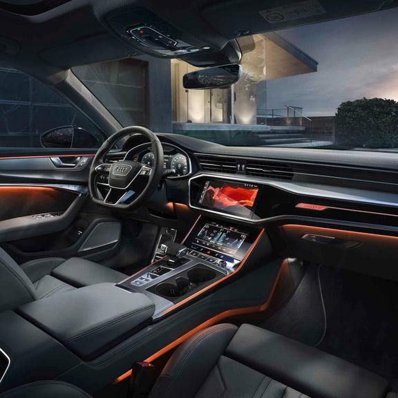 Audi Innenraum mit orangenem Ambientelicht und digitalen Displays, im Hintergrund modernes Haus mit Treppe, Hecke und Sonnenuntergang.