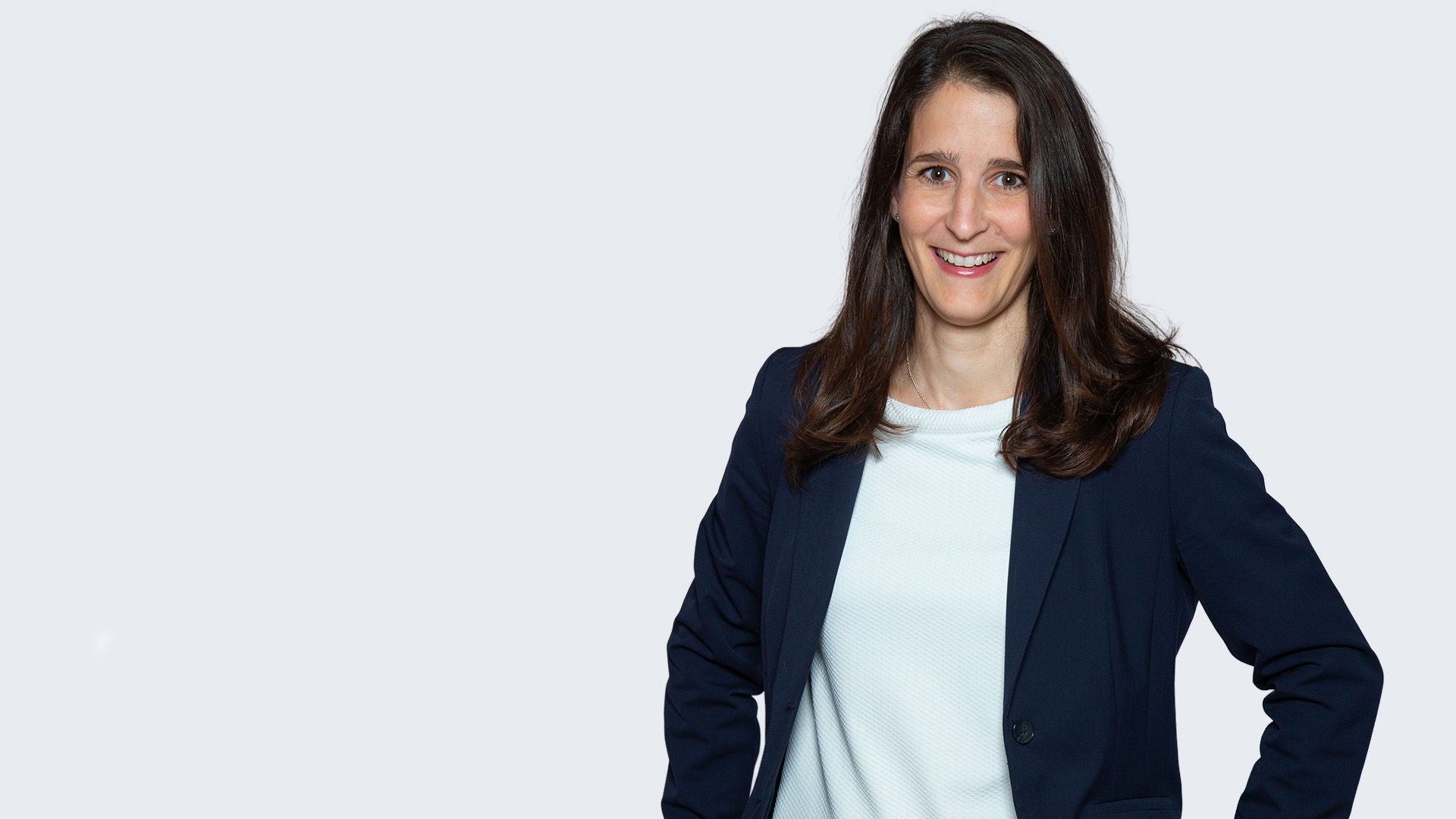 Frau im dunklen Sakko und weißem Shirt steht mit in die Hüften gestemmten Armen vor hellem Hintergrund und lächelt.