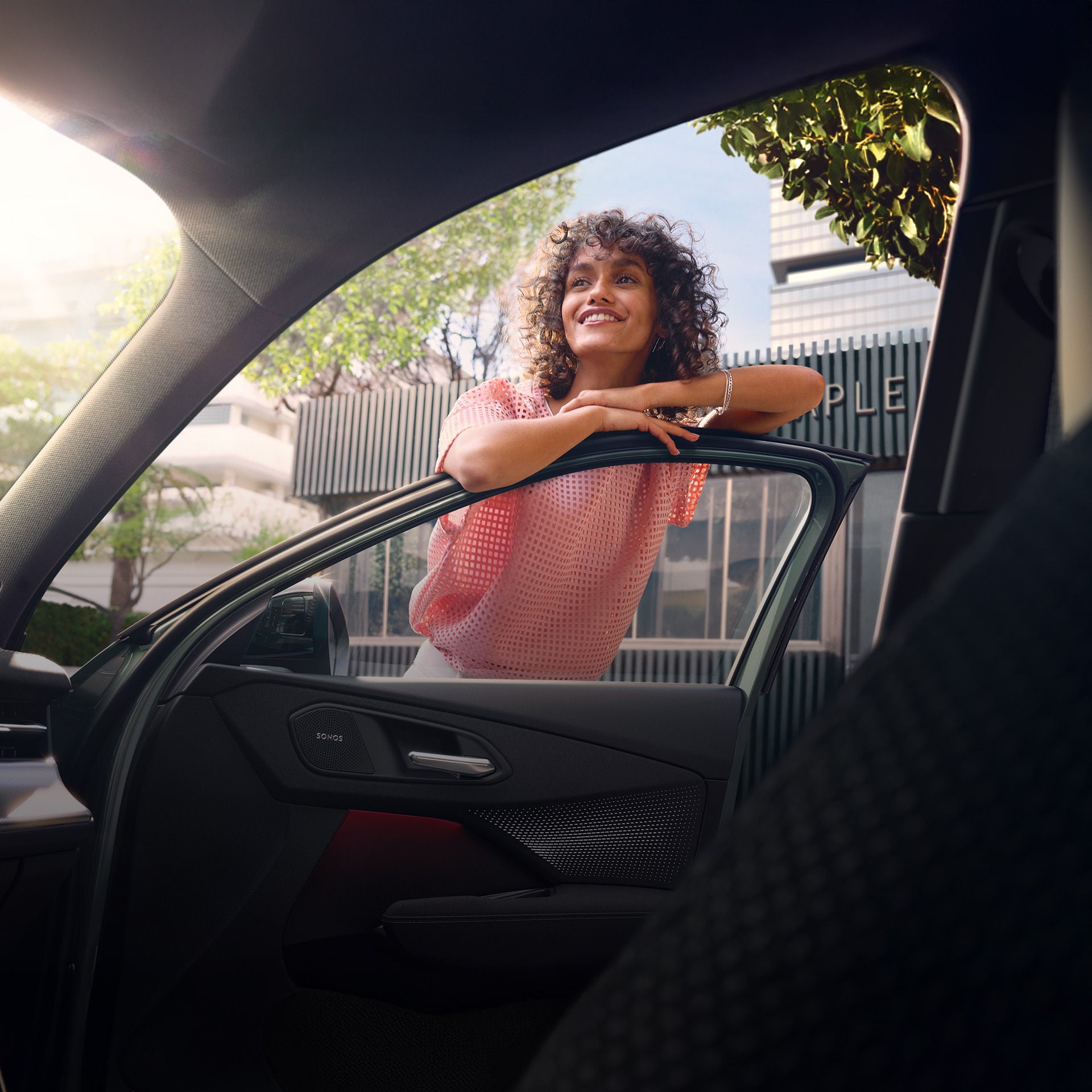 Frau lehnt fröhlich an geöffneter Autotür, Blick ins Innere mit sichtbarem Sonos-Lautsprecher und Stadt im Hintergrund.
