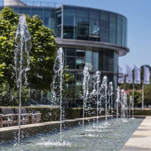 Springbrunnen vor modernem Glasgebäude an einem sonnigen Tag.