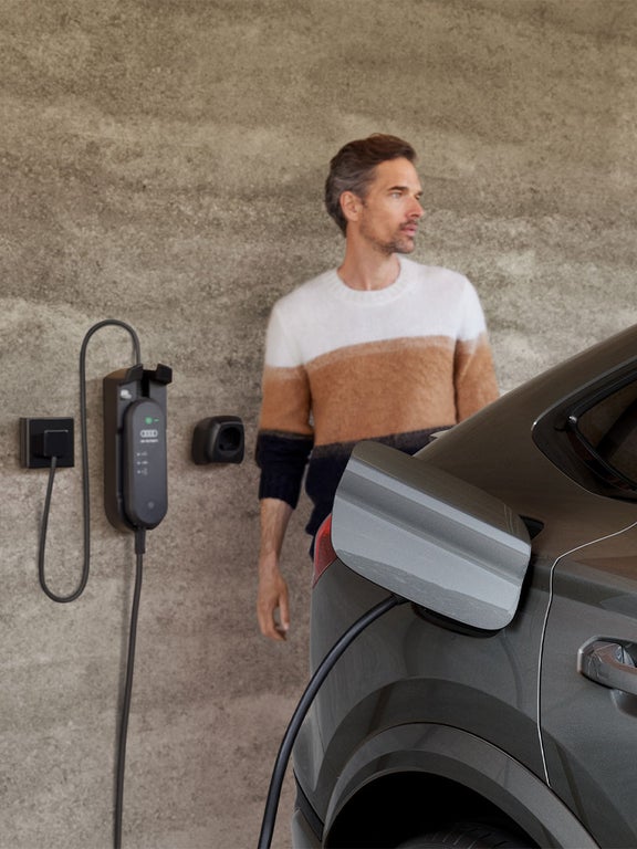 Ein grauer Audi Q6 Sportback e-tron lädt an einer Audi-Wallbox in einer Garage, ein Mann steht daneben an der Wand.