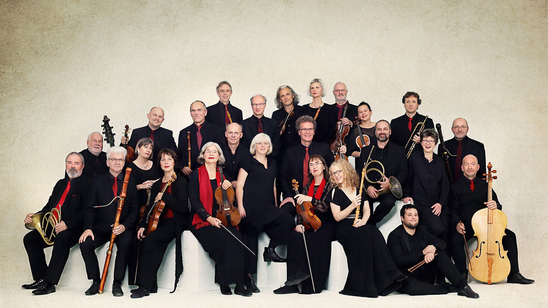 Gruppenporträt des Barockensembles Akamus: die Musikerinnen und Musiker posieren in schwarzer Konzertkleidung mit historischen Instrumenten wie Violine, Blockflöte, Horn, Trompete, Fagott und Cello vor hellem Studiohintergrund.