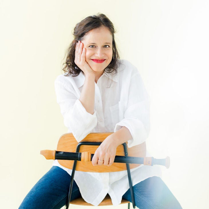 Frau mit weißem Hemd sitzt lächelnd auf einem Stuhl, hält eine Blockflöte vor hellem Hintergrund.