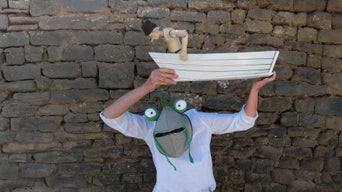Person mit Froschmaske hält eine Holzfigur in einem Boot über dem Kopf; Steinmauer im Hintergrund.