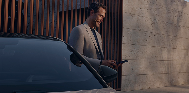 A man in a suit stands next to a sleek car, holding a phone, in front of a modern architectural backdrop.