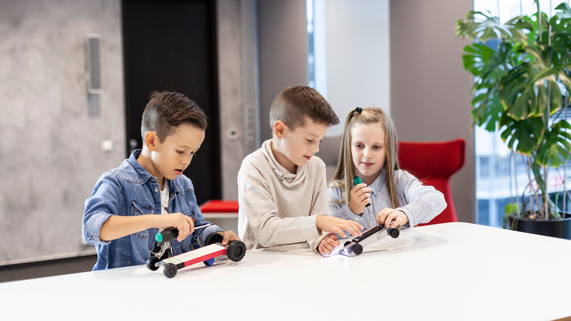Drei Kinder sitzen im Audi Forum Neckarsulm an einem Tisch und bauen kleine Rennwagen-Modelle aus verschiednen Materialien.