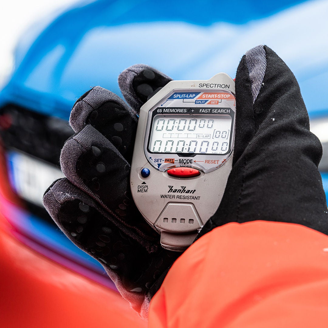 A hand wearing a black winter glove is holding a digital stopwatch; the sleeve of an orange jacket; a blurred blue-and-red background. 