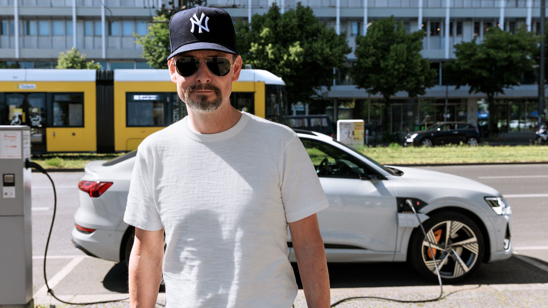 Mann mit Sonnenbrille und Baseballcap steht vor weißem Audi e-tron an Ladesäule, Straßenbahn im Hintergrund. 