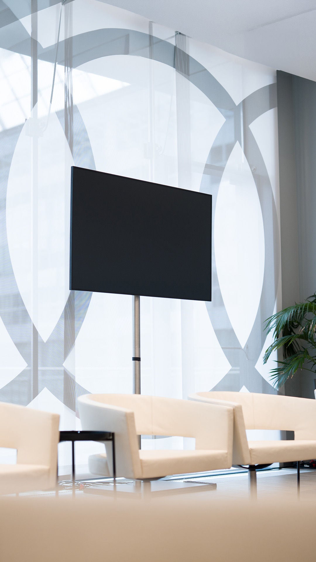 Monitor auf Stativ vor Glaswand mit Muster, helle Sessel und Pflanze im Raum.