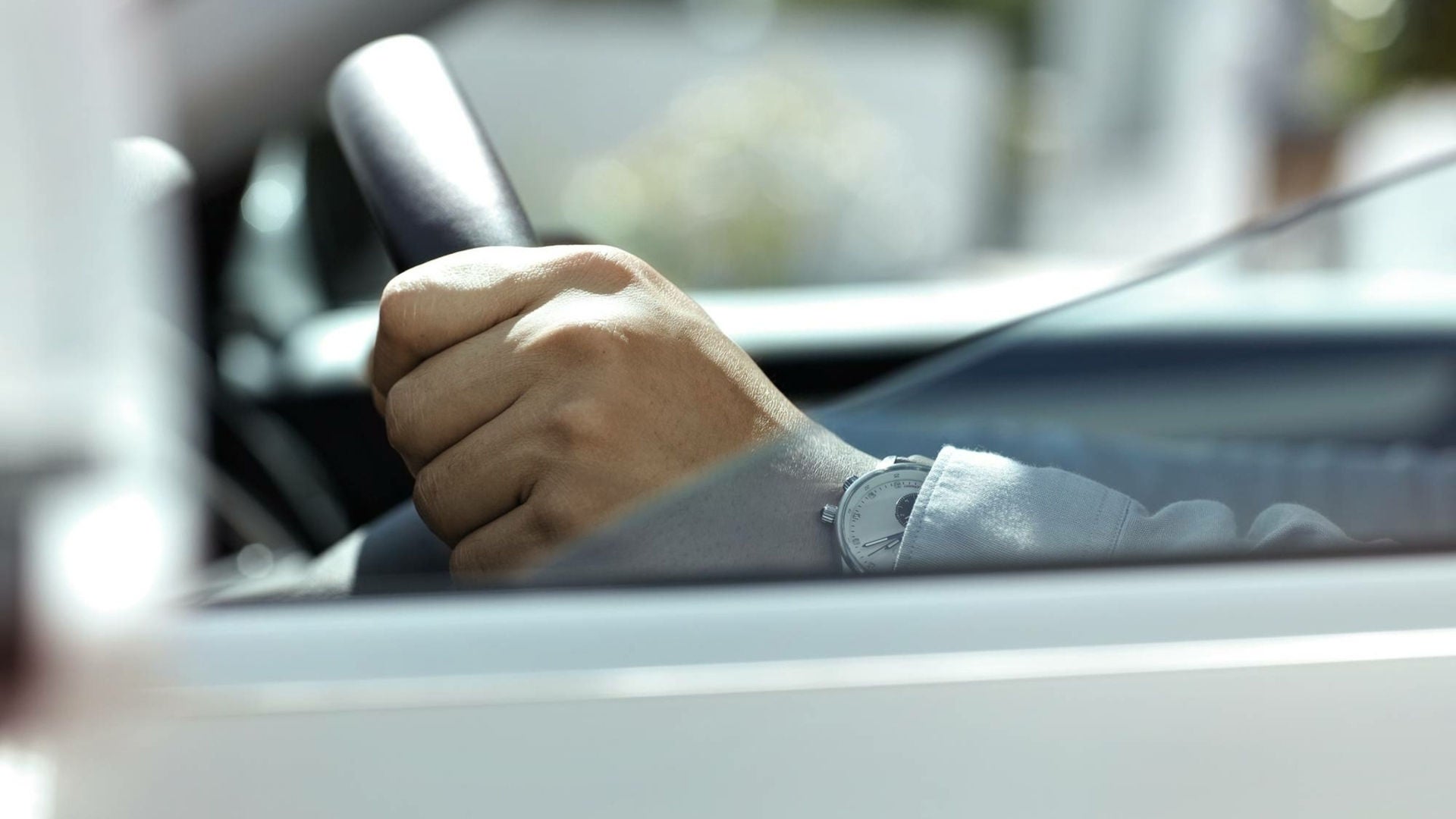 Hand am Lenkrad eines Fahrzeugs, durch das Seitenfenster fotografiert; am Handgelenk eine silberne Armbanduhr.