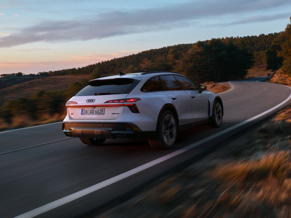The Audi A6 allroad e-hybrid, seen from the rear at an angle whilst travelling at full speed along a mountain road. A wooded area can be seen in the background.