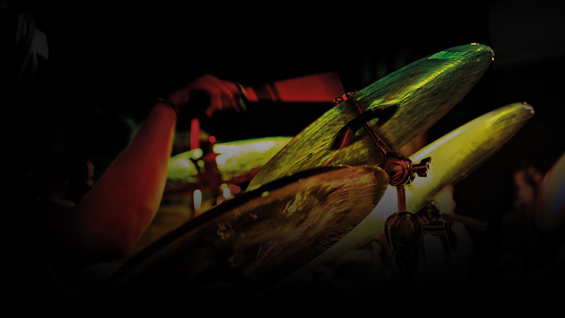 Nahaufnahme von Schlagzeugbecken und Stativ im Bühnenlicht; unscharfer Musiker im Hintergrund, grüner und gelber Lichtschein.