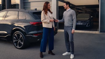 Eine Frau und ein Mann stehen vor einem Autohaus neben einem dunklen SUV vor einem Gebäude und übergeben einen Autoschlüssel. Im geöffneten Eingang ist ein weiteres Audi Modell zu sehen.